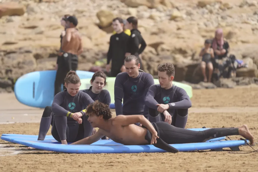 Beginner surfers sitting around an instructor lying on a board on the beach, learning the basics in Taghazout