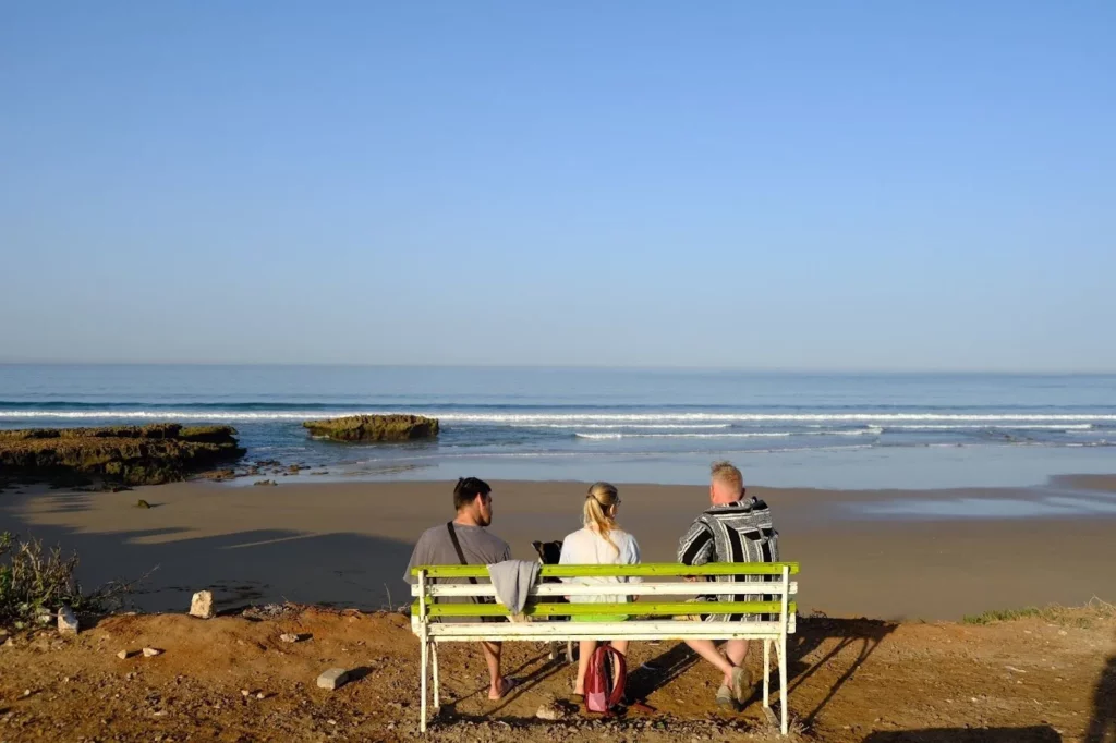 Drei Personen auf einer Bank mit Blick auf den Strand von Tamraght und das Meer